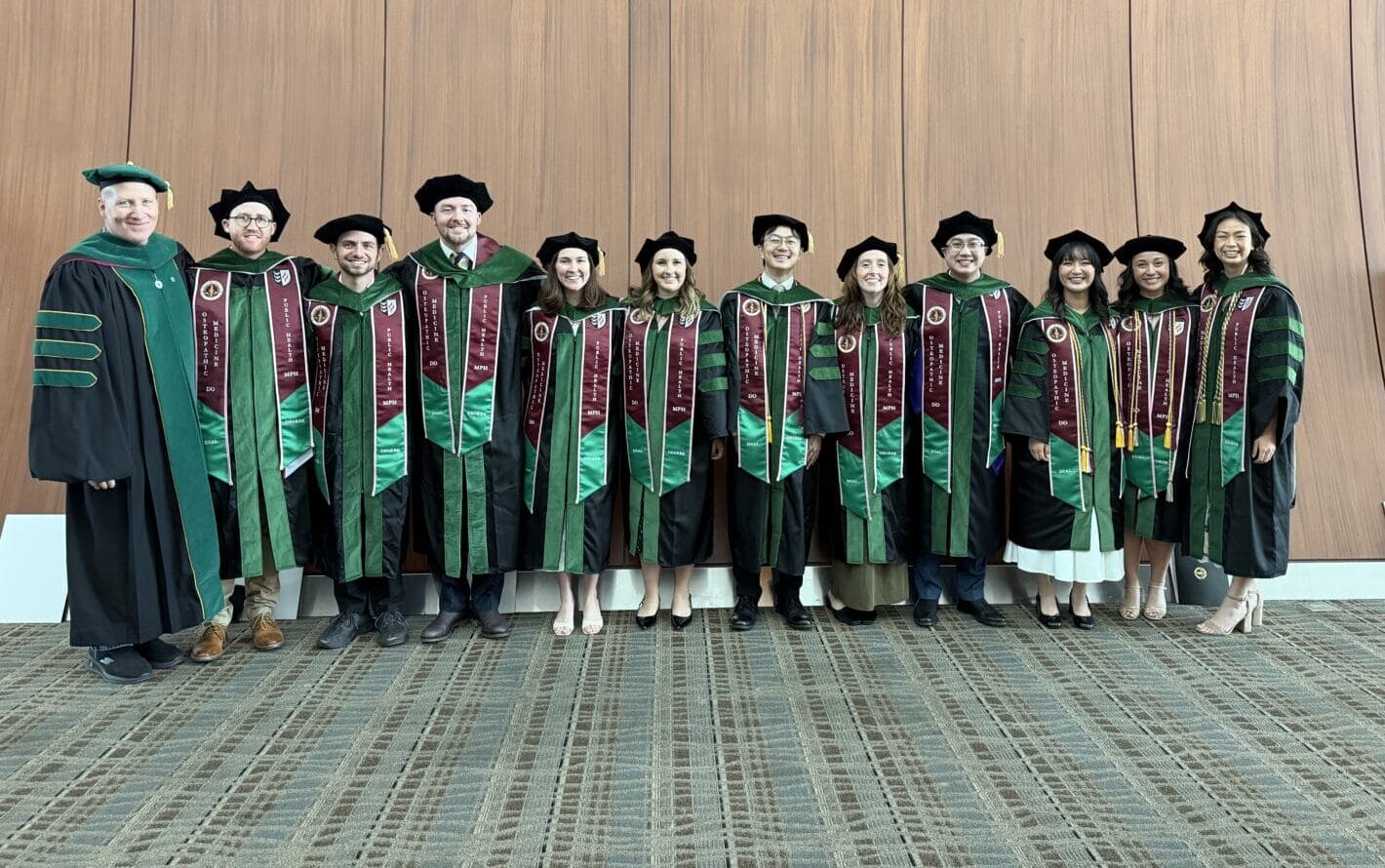 A group of eleven graduates in green academic robes and hats stand in a row indoors with a faculty member, posing for a photo against a wood-paneled wall.