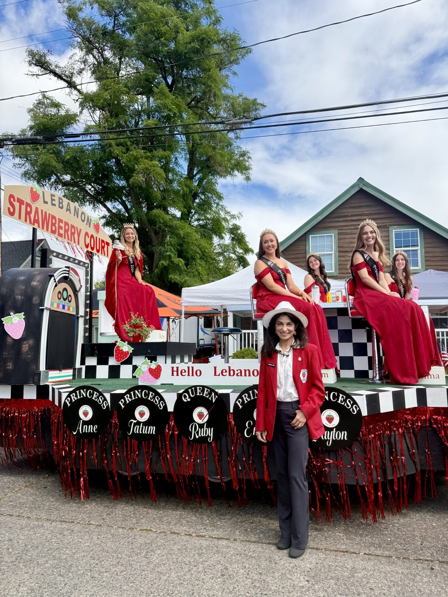 Five women in red gowns sit on a parade float labeled "Lebanon Strawberry Court," with another woman in a red jacket standing in front on the street, during a community event.