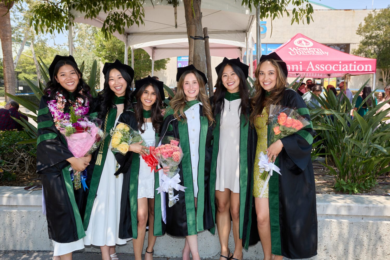 Six women in graduation gowns and caps stand outdoors, holding flowers and smiling for a group photo. A sign reading "Alumni Association" is visible in the background.