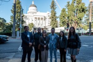 Six people stand and pose for a group photo on a street with a white domed government building and tall trees in the background.