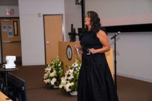 A woman in a black dress speaks at the front of a lecture room with a microphone. Floral arrangements and presentation equipment are visible in the background.