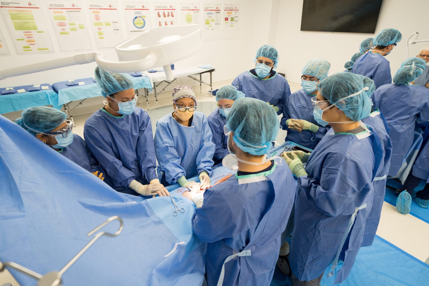 Medical team in blue scrubs and masks performs a procedure on a patient in a brightly lit operating room, while several others observe and assist. Charts and equipment are visible in the background.