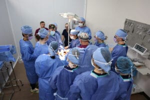A group of medical professionals in scrubs and masks gather around a patient on an operating table in a brightly lit surgery room.