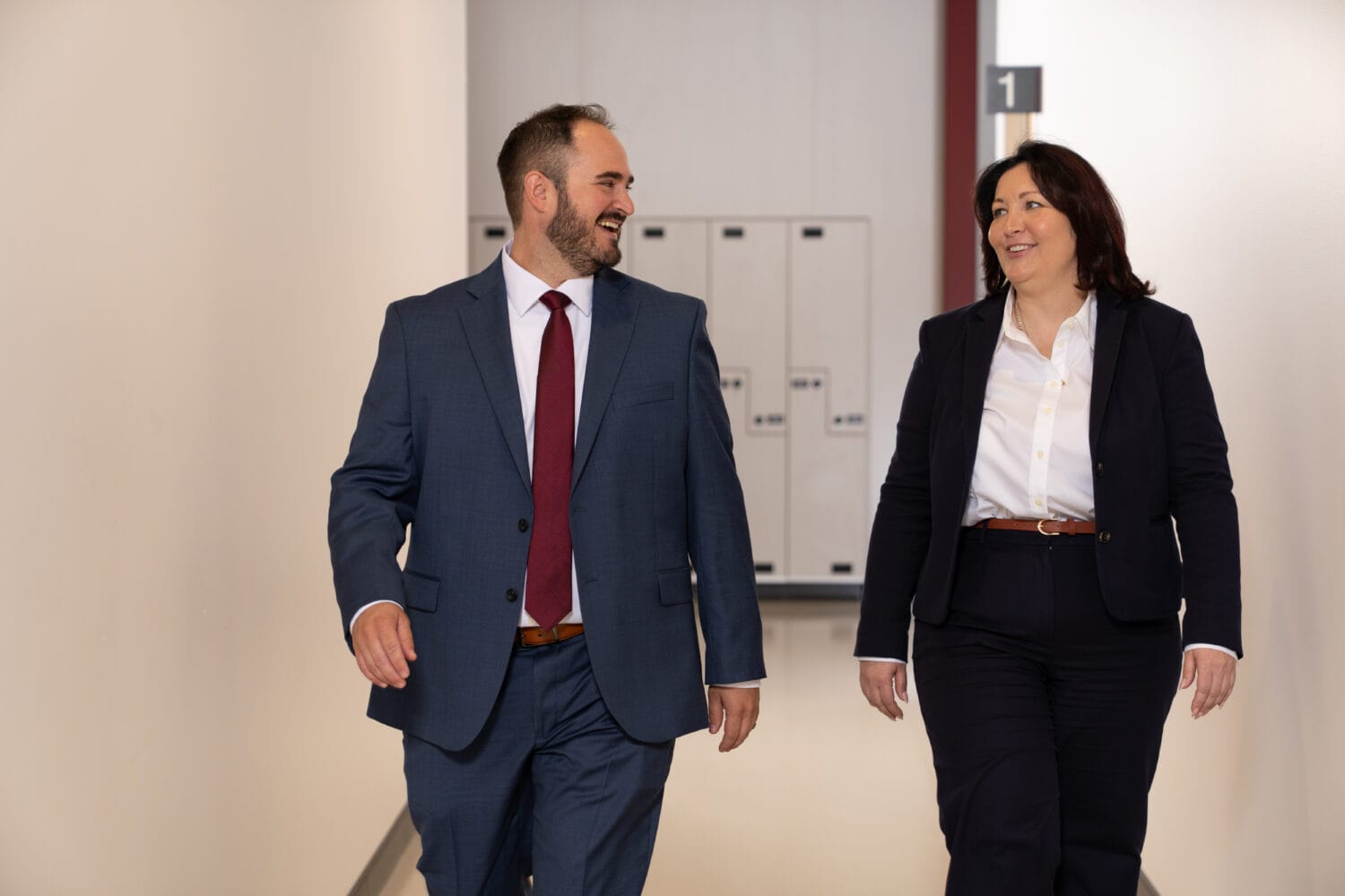 Two people in business suits walk down a hallway, talking and smiling, with lockers and a room number visible in the background.