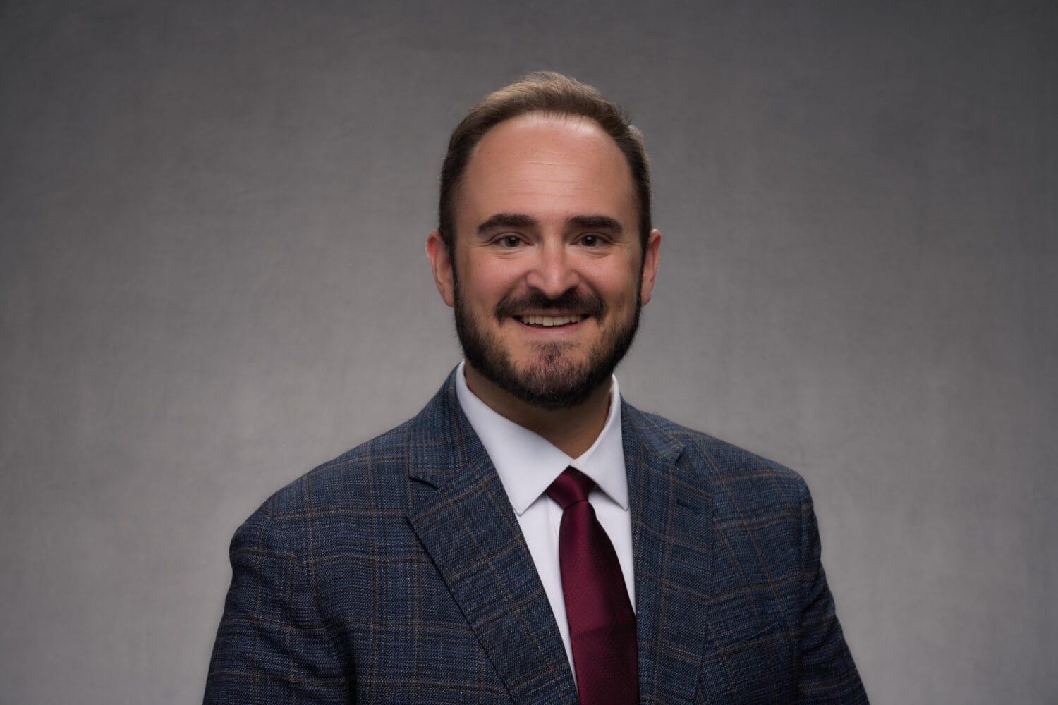 Man in a checked suit jacket, white shirt, and maroon tie smiling in front of a plain gray background.