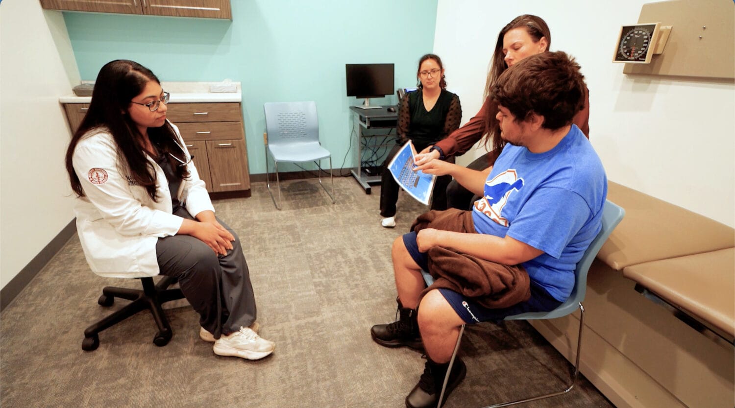 A doctor sits facing a patient and two companions in a medical exam room, as one companion shows the patient a document or image.