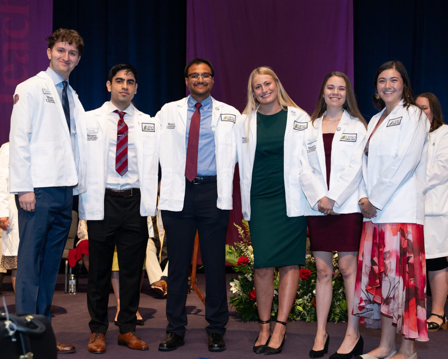 Six people wearing white coats stand together and smile on stage during a ceremony, with others seated in the background.