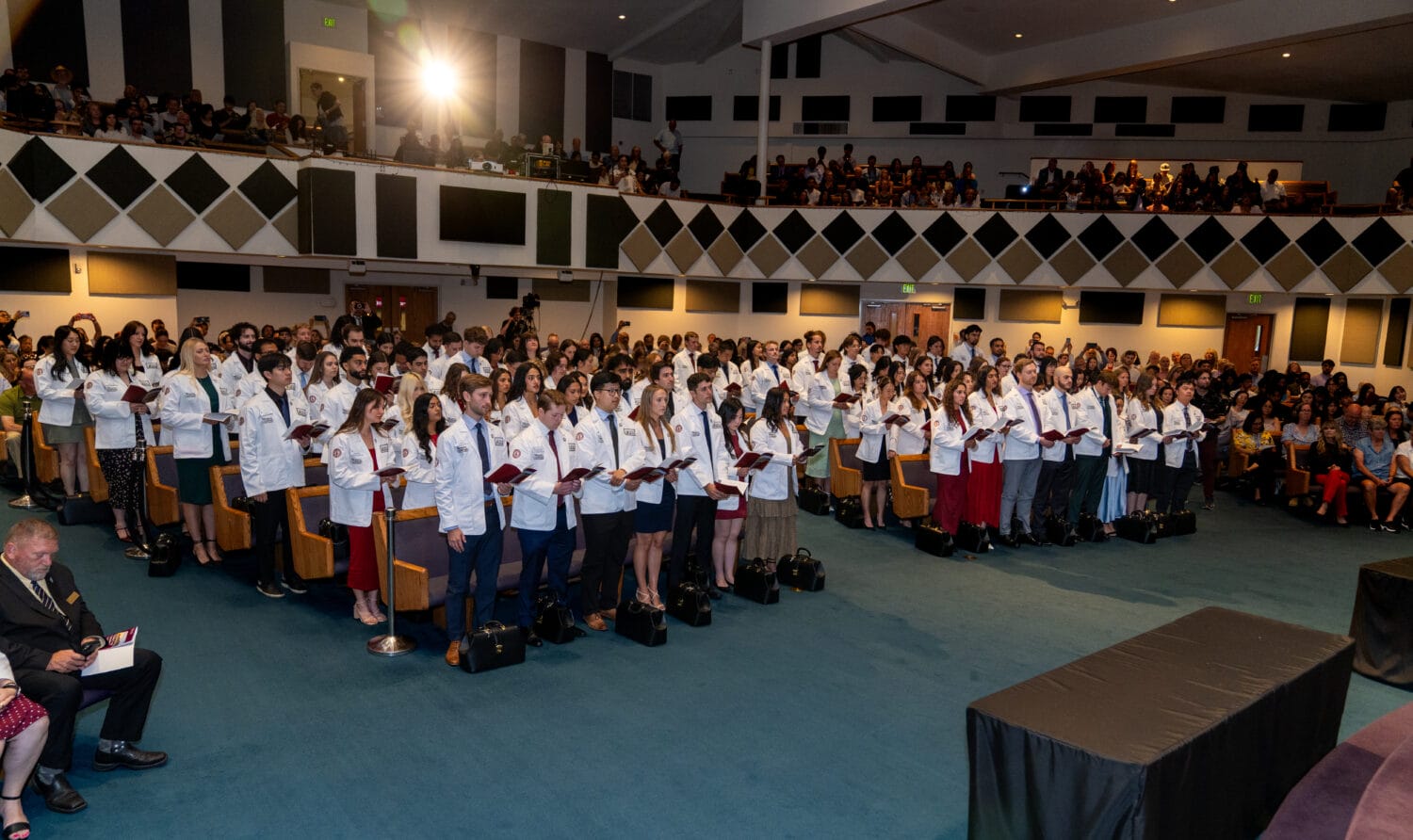 A large group of people in white coats stand in rows at a ceremony in an auditorium, holding papers, while an audience watches from the seats and balcony.