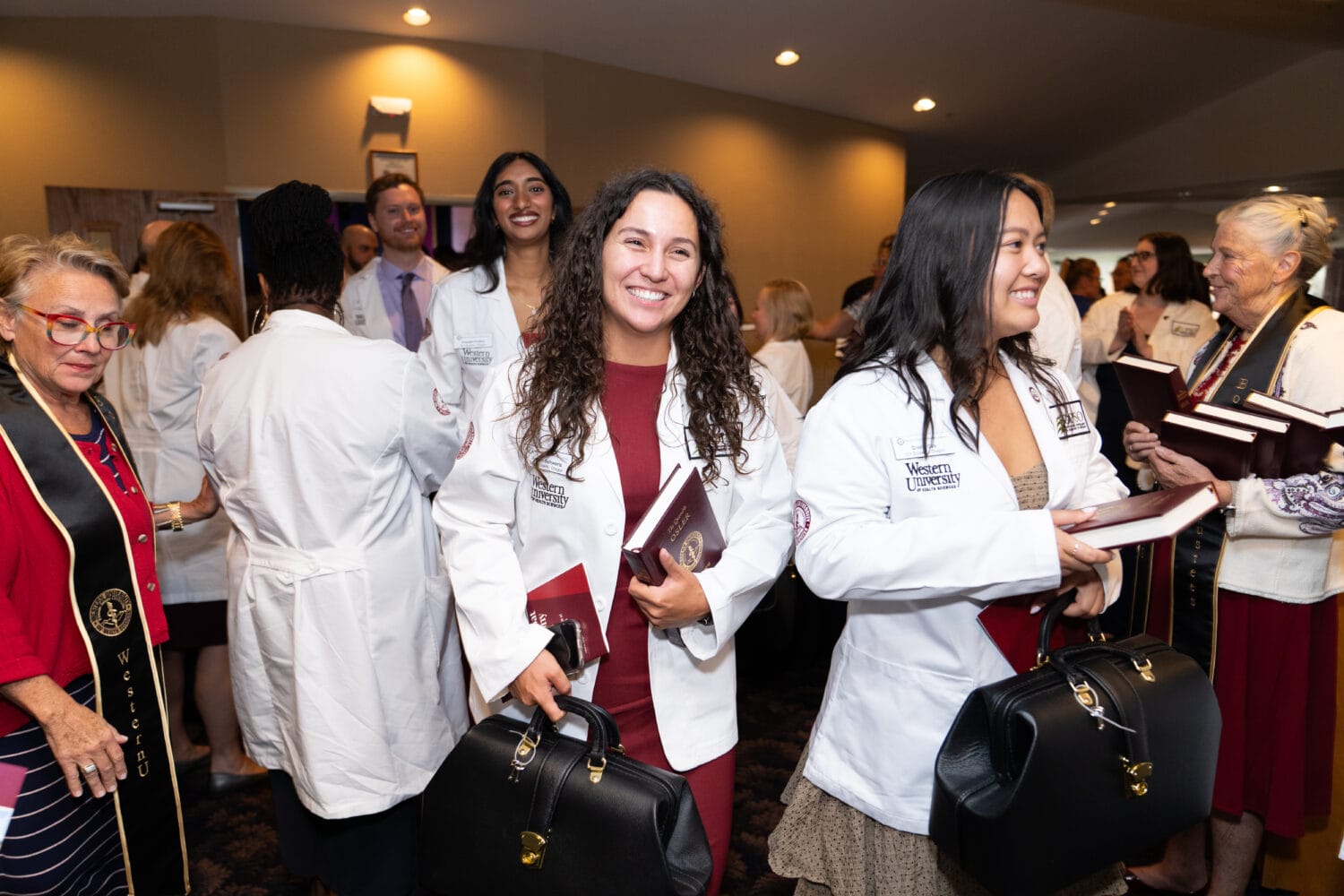 A group of people in white lab coats stand together indoors, some smiling and holding black bags and books at what appears to be a professional or academic event.