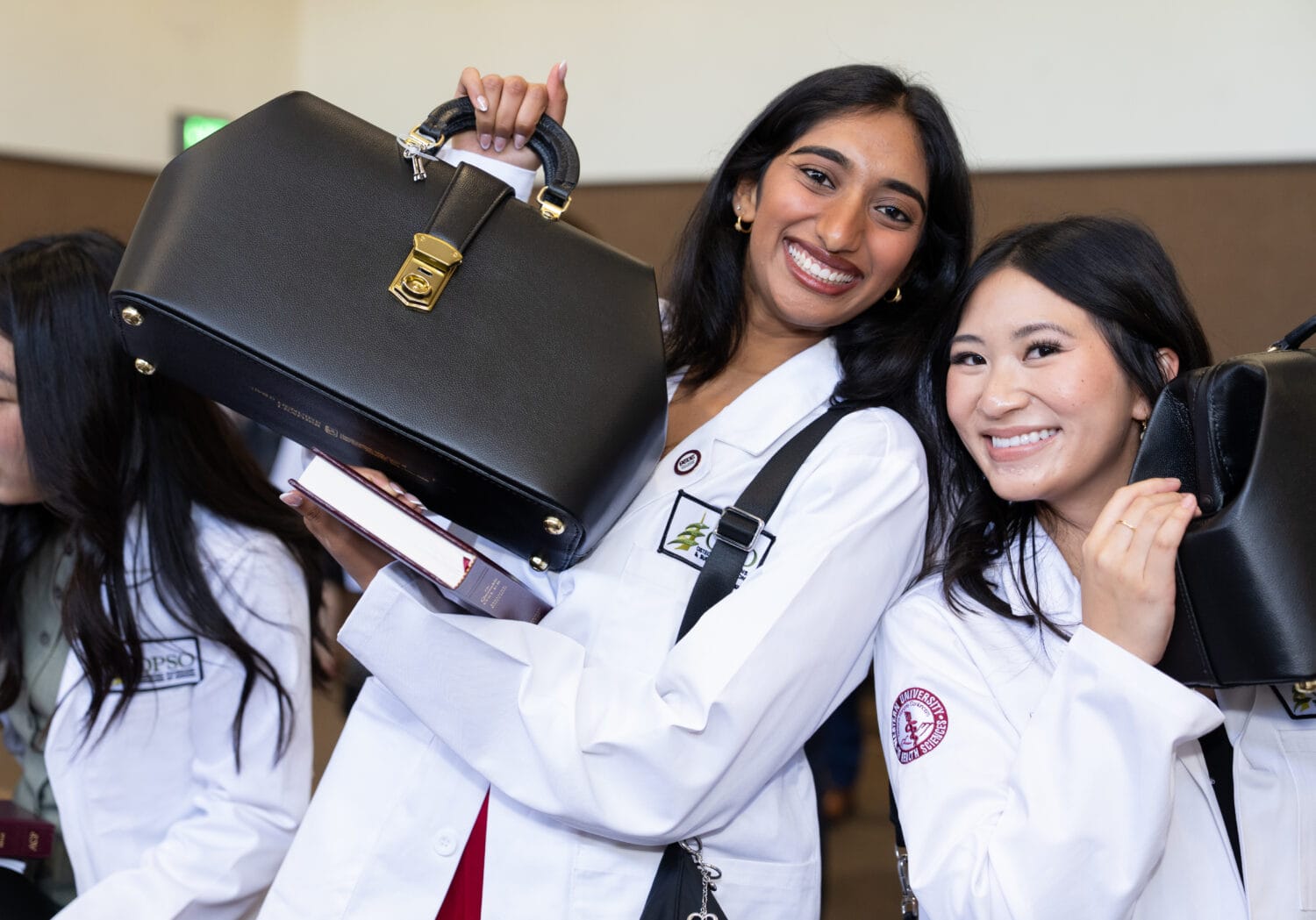 Two women in white coats smile and hold black medical bags; another person is partially visible to the left.