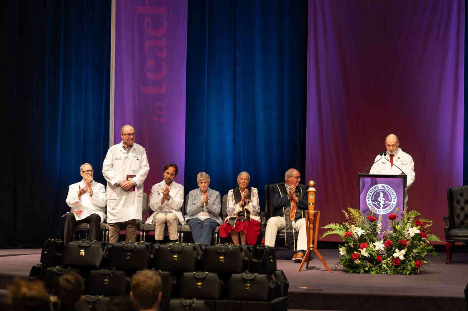 A group of people in white coats and formal attire sit and stand on stage at a ceremony, with one person speaking at a podium beside a flower arrangement.