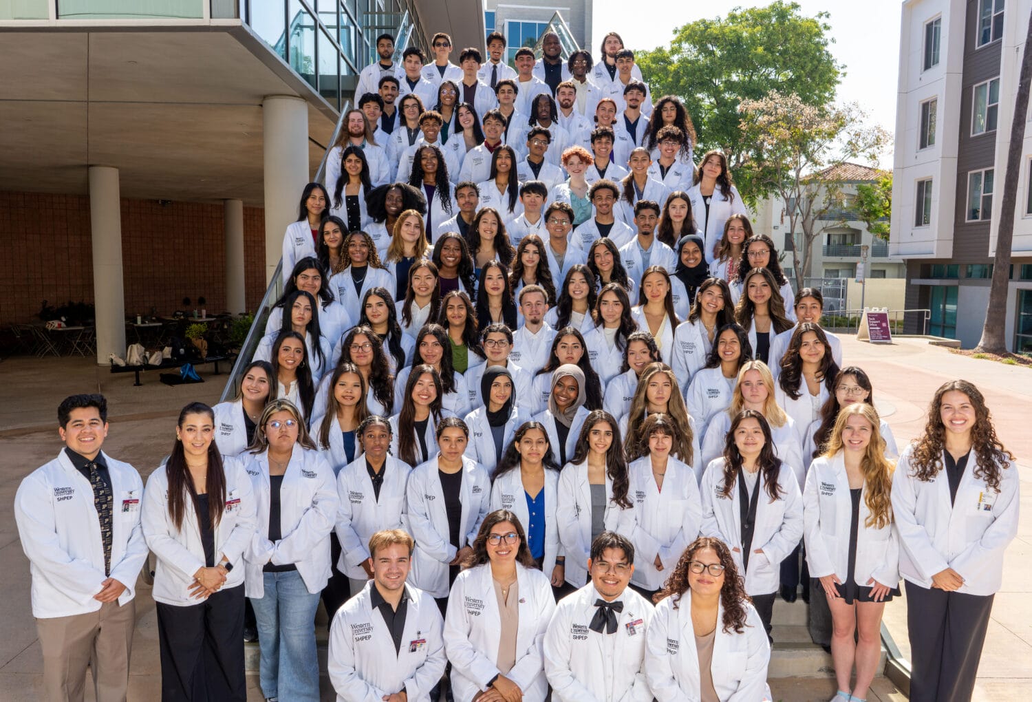 A large group of students in white coats poses together on outdoor steps in front of a campus building.