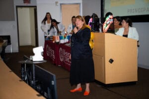 A woman speaks at the front of a lecture hall with attendees and a table covered with maroon tablecloths displaying promotional items in the background.