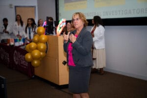 A woman in a suit speaks to an audience in a classroom, with people in white coats and yellow balloons in the background near a presentation screen.