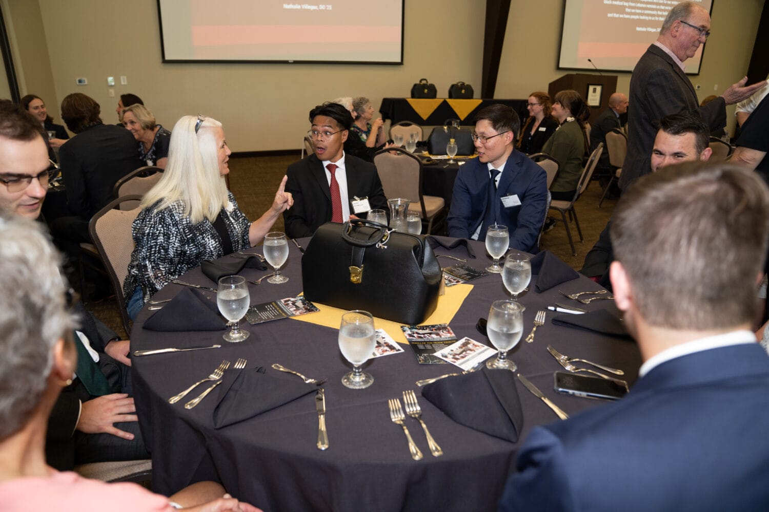 People seated around a round table with black napkins at a formal event, conversing before a presentation; a large black bag is on the table.
