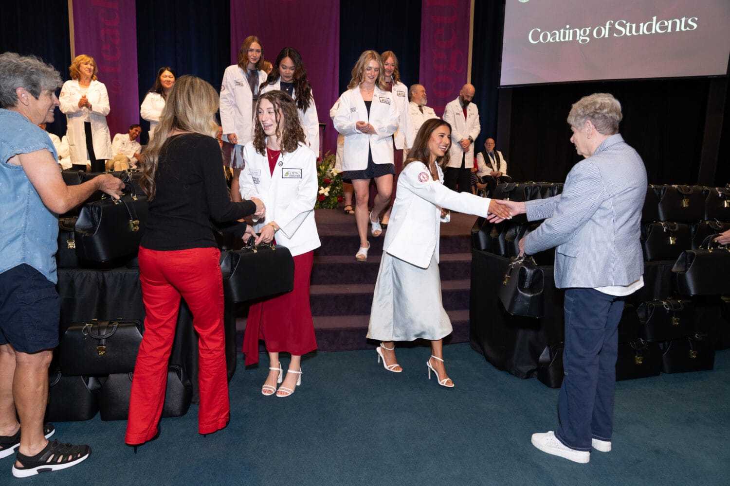 Students in white coats receive black bags and shake hands with presenters on stage during a ceremony, while others wait in line and observe.
