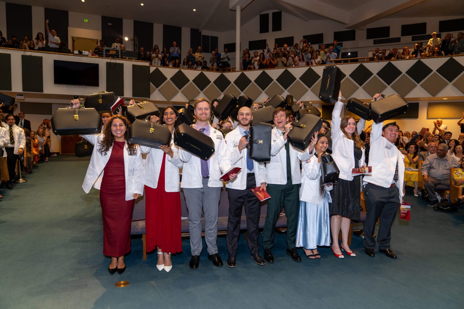 A group of people in white coats stand in front of an audience, holding up black cases and smiling, as part of a ceremony in a large indoor auditorium.
