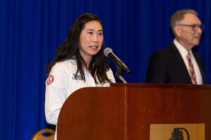 A woman in a white coat speaks at a podium with a microphone; a man in a suit stands in the background against a blue curtain.