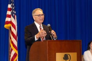 A man in a suit speaks at a podium with a microphone. An American flag and a blue curtain are in the background.