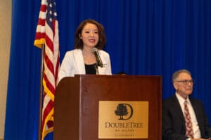 Woman in a white blazer speaks at a podium labeled “DoubleTree by Hilton Claremont” with an American flag and a seated man in the background.