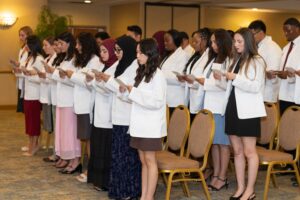 A group of people wearing white coats stand in a row, reading from papers during a ceremony in a carpeted room with chairs.