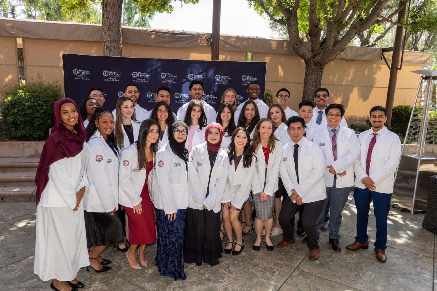 A group of students in white coats pose for a photo outdoors in front of a backdrop with university logos and trees.