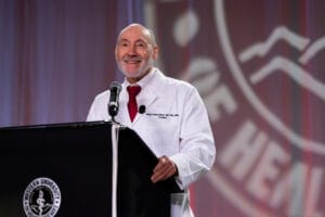 An older man in a white lab coat stands at a podium with a microphone, smiling, in front of a blurred university emblem backdrop.
