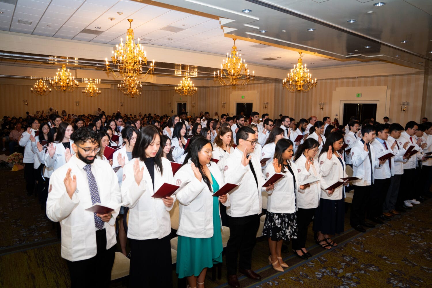 A group of medical students in white coats stands with raised right hands, reciting an oath at a ceremony in a chandelier-lit banquet hall.
