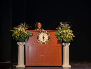 A woman stands at a podium with the Western University of Health Sciences seal, flanked by two floral arrangements on pillars, against a dark background.
