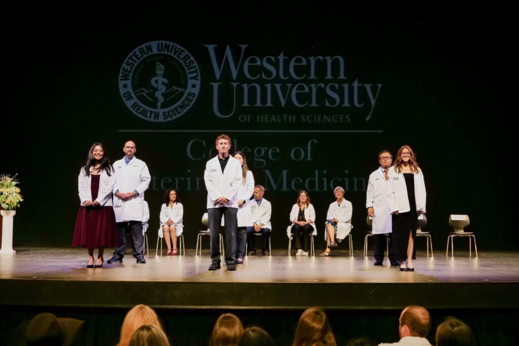 A group of people wearing white coats stands on stage at a Western University of Health Sciences event, with others seated behind them.