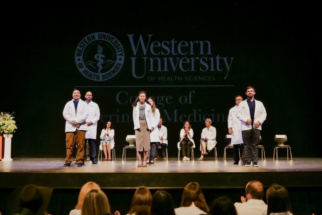 Seven people in white coats stand on stage, with others seated behind them and the Western University of Health Sciences logo projected in the background.
