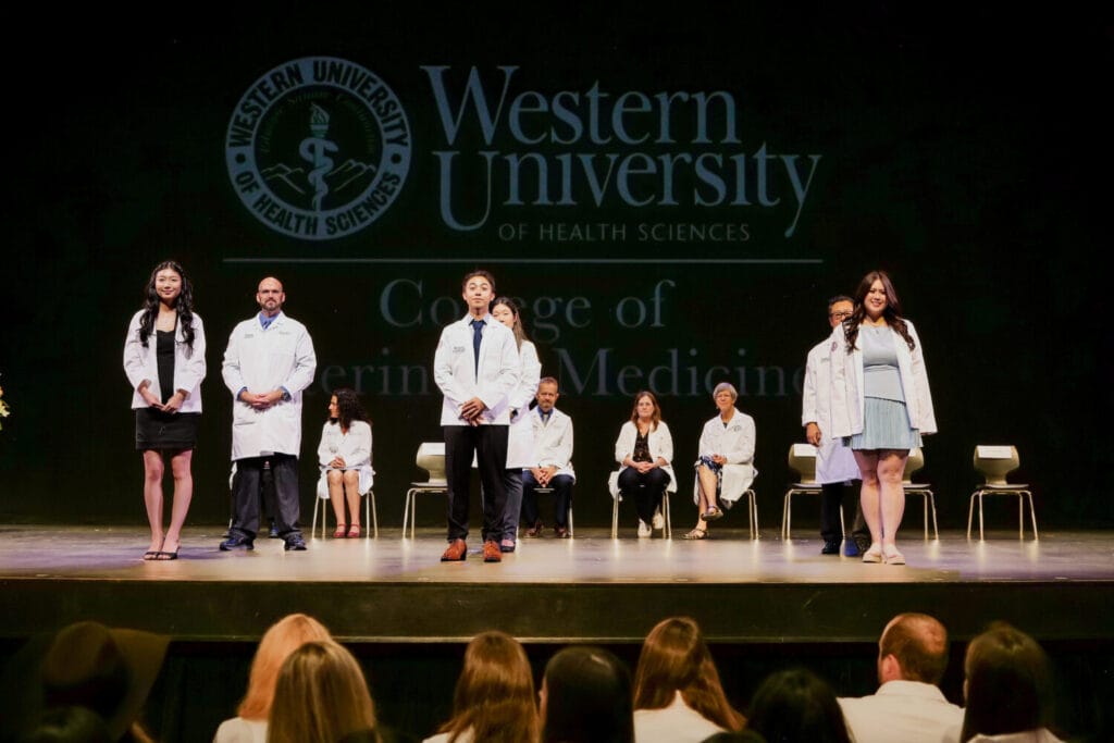 Medical students in white coats stand on stage at a Western University of Health Sciences ceremony, with officials and faculty seated behind them and an audience in the foreground.