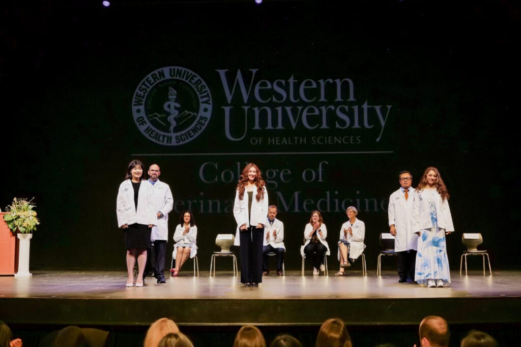Several people in white coats stand on stage at a Western University of Health Sciences event, with others seated behind them and a podium with flowers on the left.