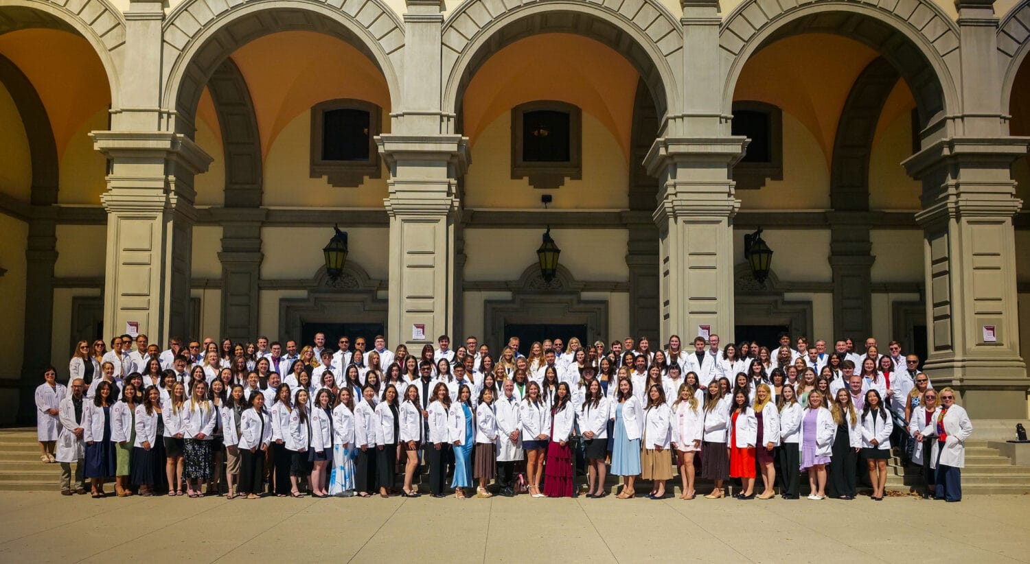 A large group of people in white coats poses together outside a building with tall arches and stone columns.