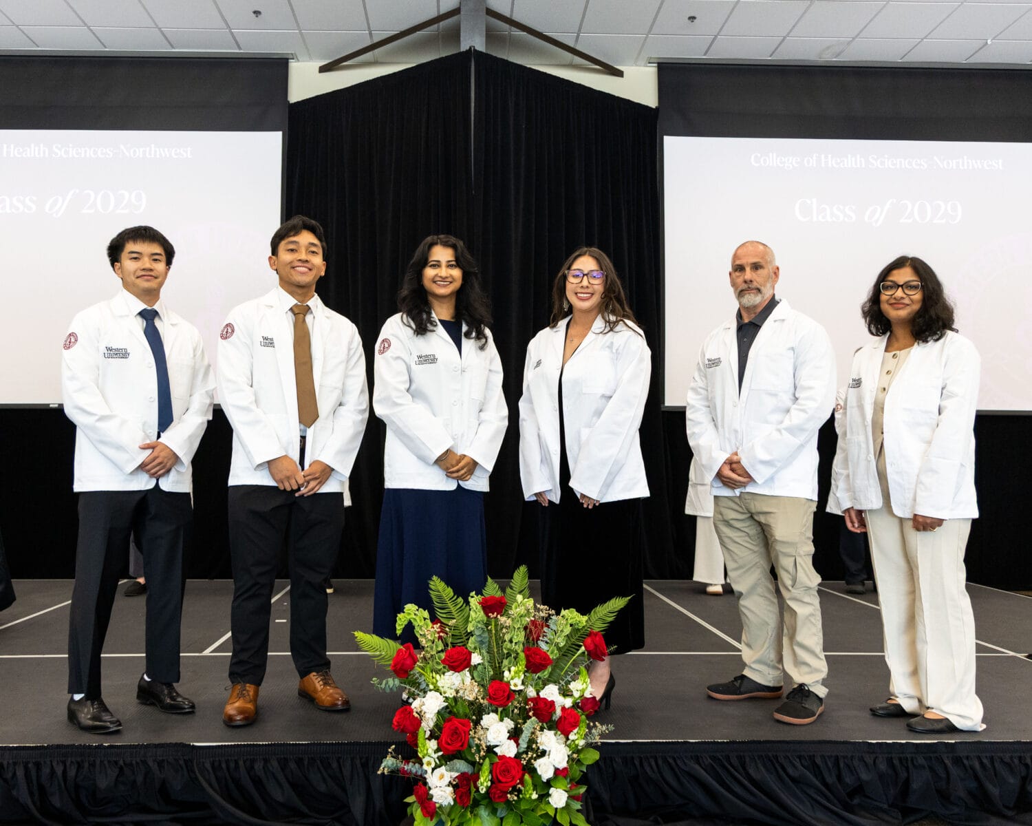 Six people in white coats stand on stage in front of black curtains, with a red and white flower arrangement in front of them and Class of 2029 signs in the background.