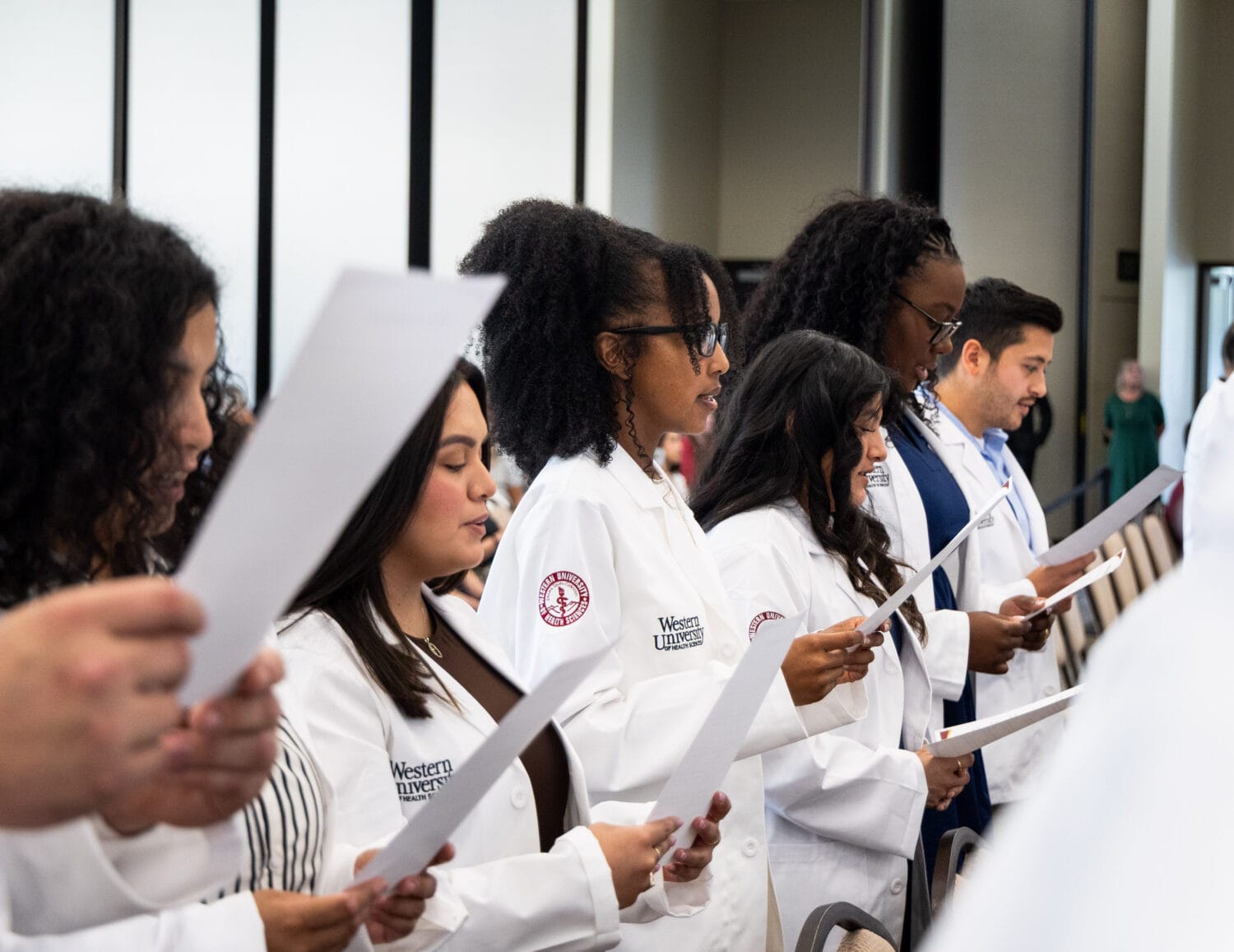 A group of students in white coats stands in a row, holding papers and reading aloud during an indoor ceremony.