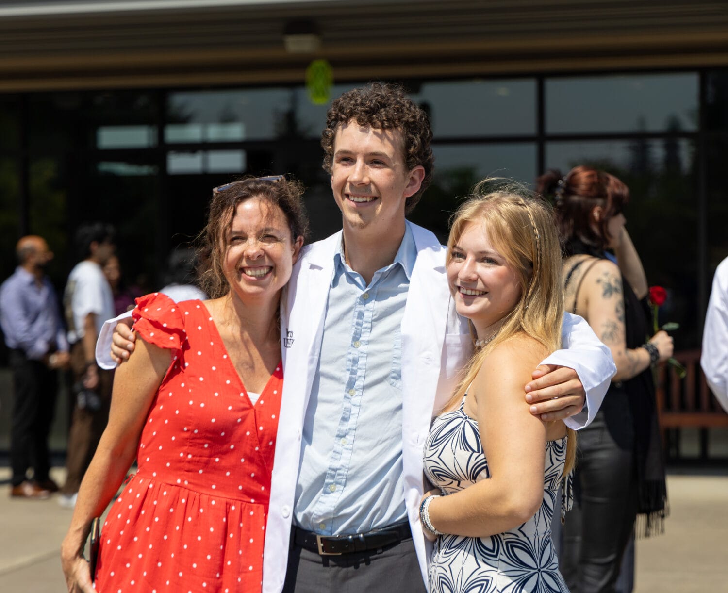 Three people smiling and posing together outdoors, with one wearing a white coat. Other people are visible in the background.