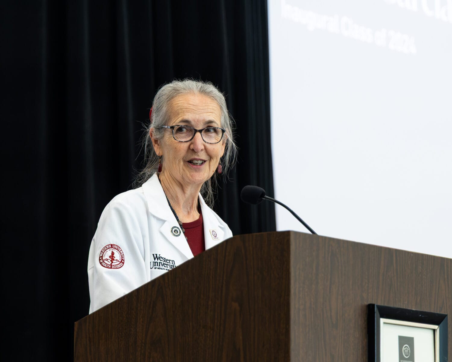 An older woman in a white lab coat speaks at a podium with a microphone. A projection screen with partial text is visible in the background.