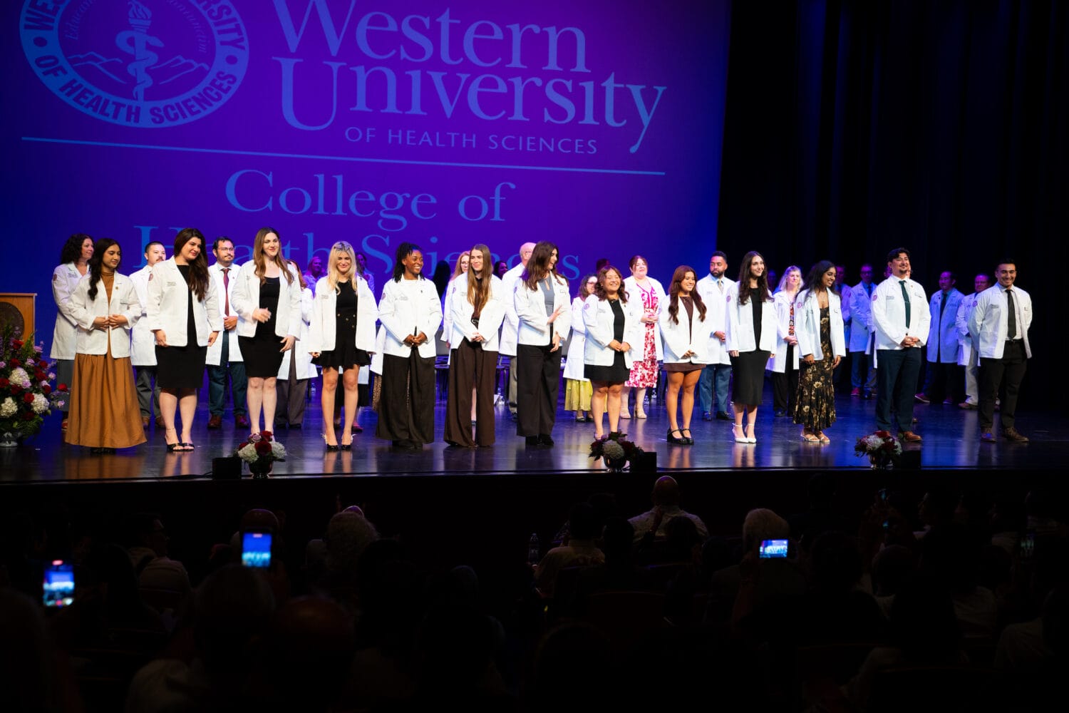 A group of people in white coats stand on stage at the Western University College of Health Sciences ceremony, facing an audience.