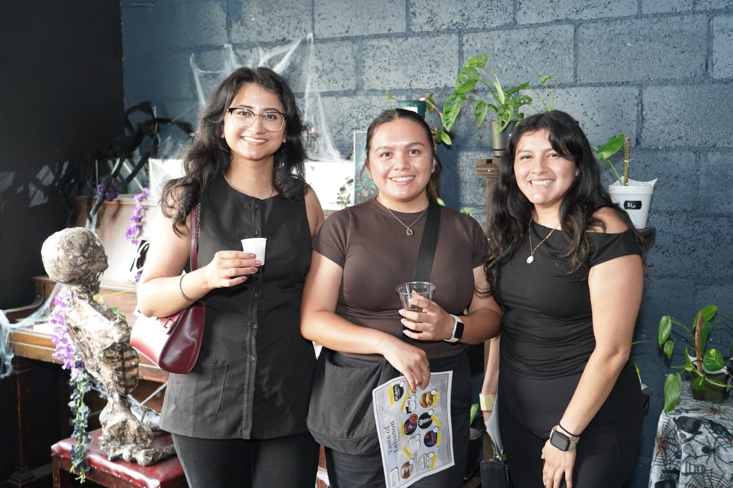 Three women stand indoors, smiling at the camera, each holding a drink. Plants and Halloween-themed decorations, including cobwebs and a skull, are visible in the background.