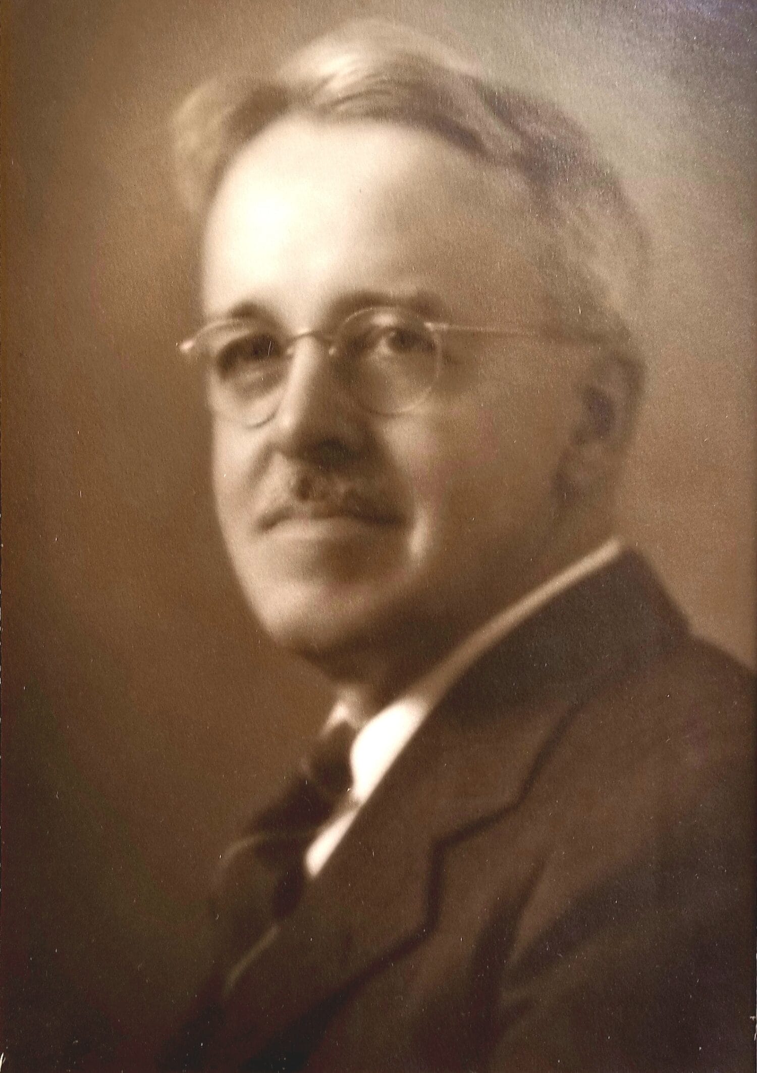 Sepia-toned portrait of an older man with glasses, a mustache, and a suit, looking slightly to the side.