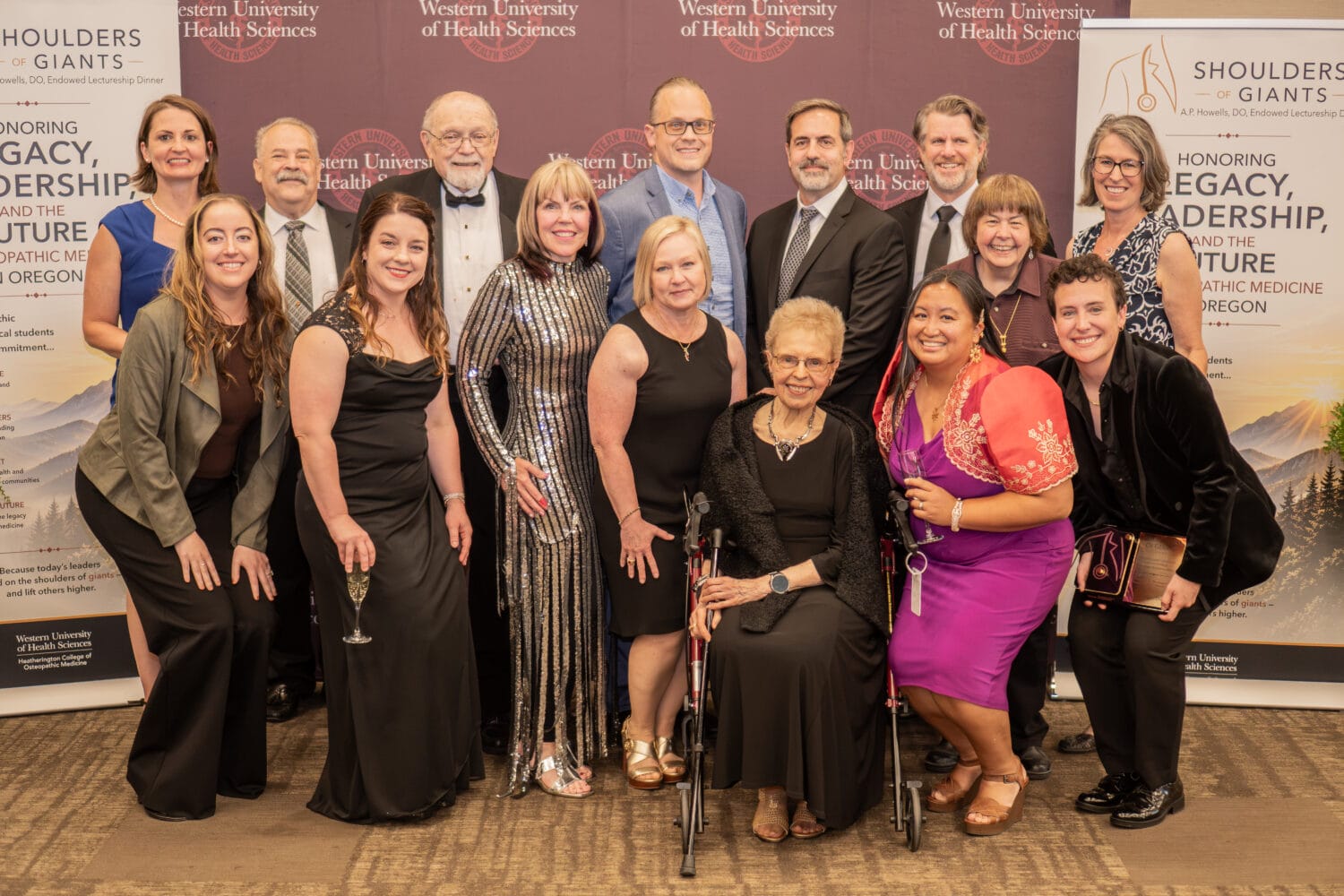 A group of people dressed formally pose together at a Western University of Health Sciences event with a banner reading “Shoulders of Giants” in the background.