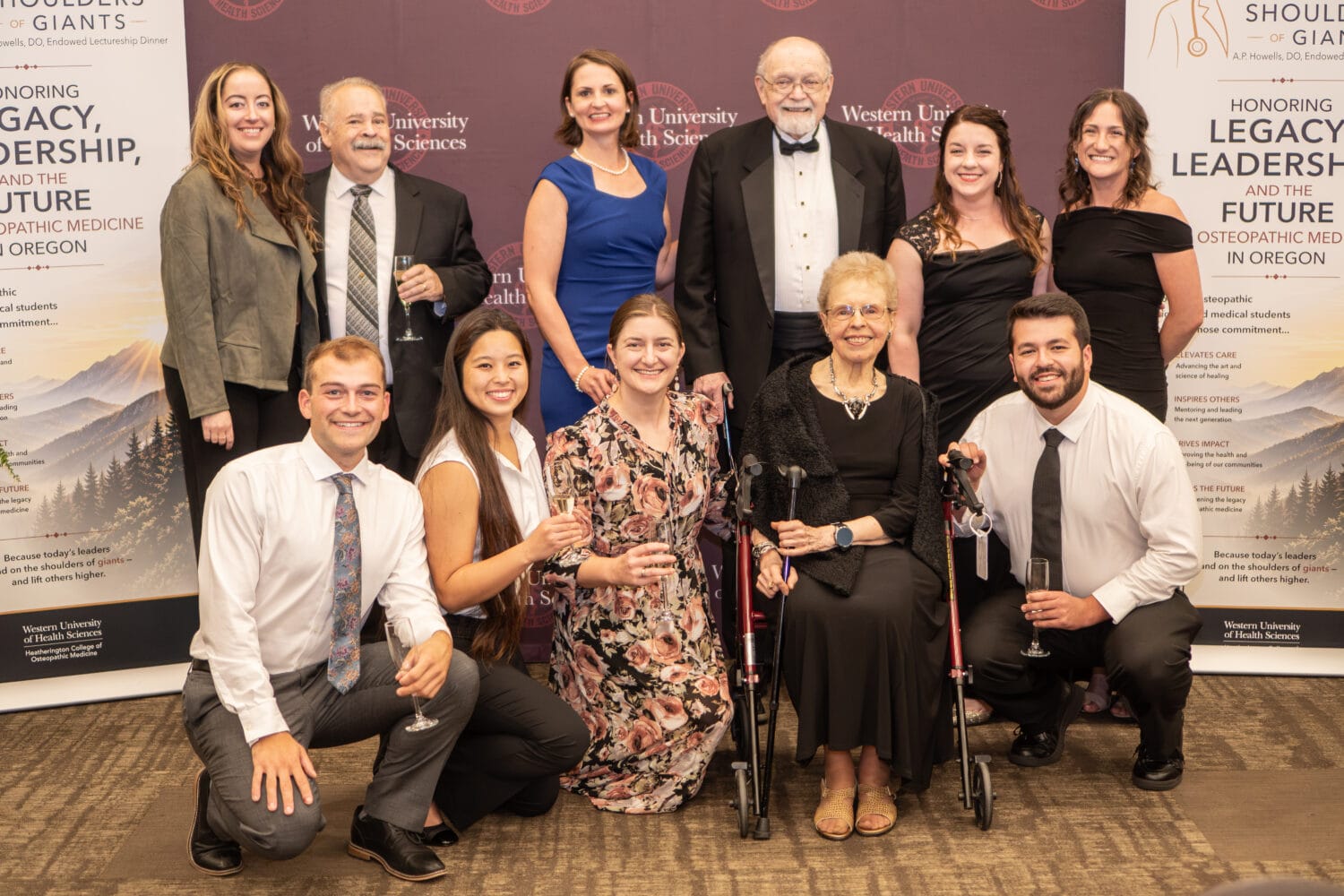 A group of people, dressed formally, pose together at an indoor event. Two banners about legacy and leadership in osteopathic medicine are visible in the background.