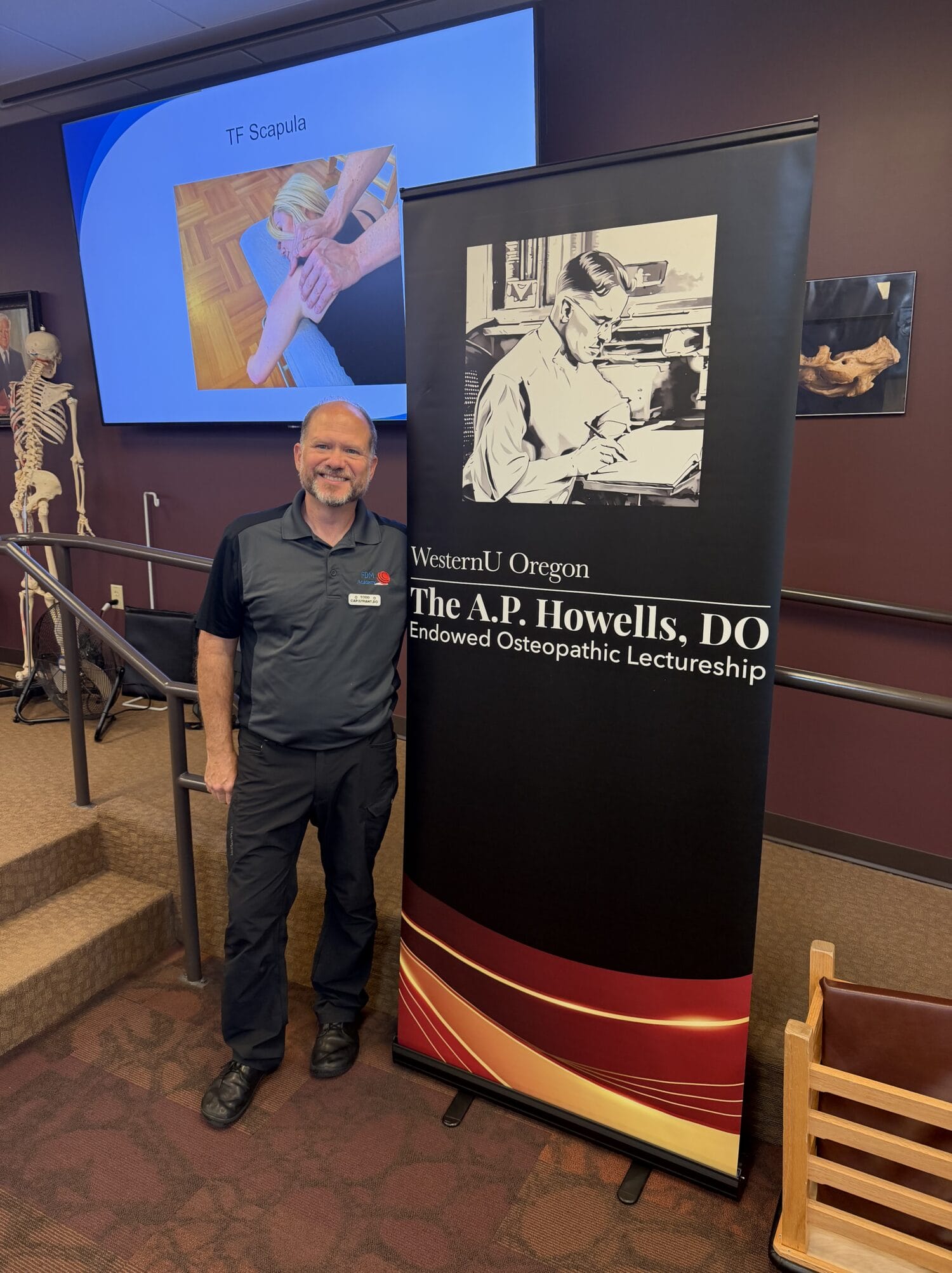 A person stands beside a WesternU Oregon A.P. Howells, DO, Endowed Osteopathic Lectureship banner in a classroom with a medical presentation and skeleton model visible in the background.
