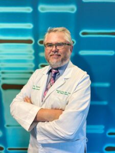 A man in a white lab coat stands with arms crossed in front of a blue background with DNA patterns. He wears glasses, a tie, and has gray hair and a beard.