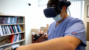 A person in blue scrubs and a face mask uses a VR headset and handheld controller.