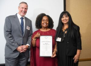 Three individuals pose together at an event, with the central person holding a Certificate of Recognition.