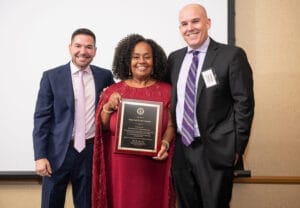 Three individuals pose together at an event, with the central person holding a framed award plaque.