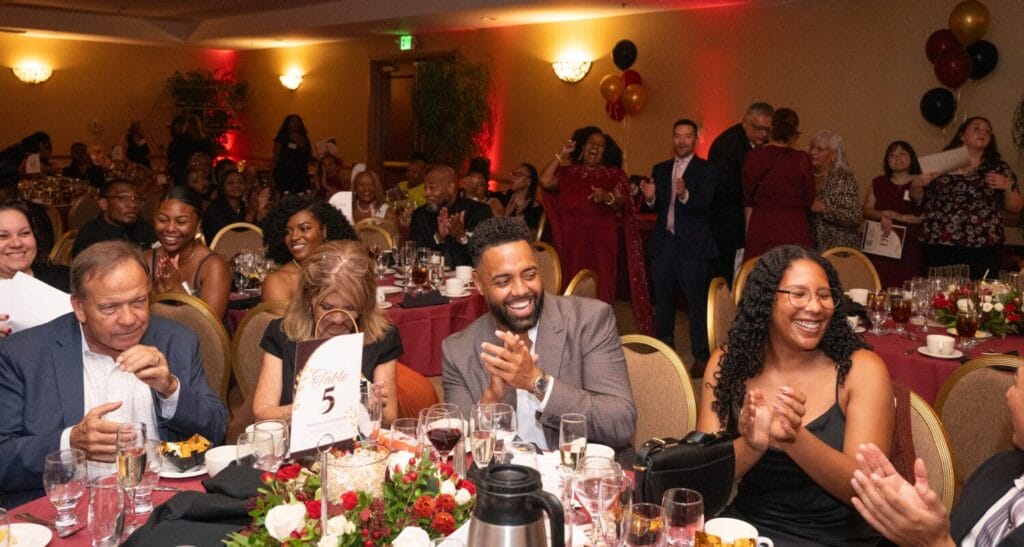 A group of people at a formal dinner event are clapping and smiling, surrounded by tables set with food, drinks, and floral arrangements.
