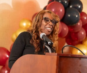 A person with long curly hair and glasses smiles while speaking into a microphone at a podium, with colorful balloons in the background.